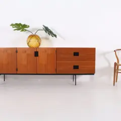 Buffet en teck par Cees Braakman, avec un vase en verre texturé et une feuille de Monstera. Une chaise Wishbone est placée à droite.