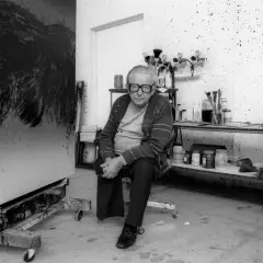 Hartung, peintre, pose dans son atelier, devant une de ses toiles abstraites. L'espace est encombré de matériel artistique.