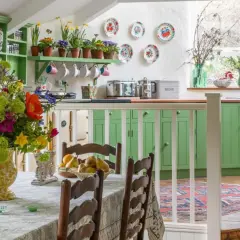 Une cuisine lumineuse avec des armoires vertes vives, un grand bouquet de fleurs colorées sur la table, et une décoration style cottage ou campagne chic.