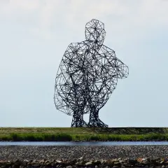 Sculpture d'Antony Gormley, une silhouette humaine filaire accroupie, se dresse sur une digue herbeuse, surplombant un canal.
