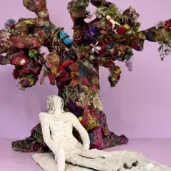 Sculpture d'arbre colorée et texturée, évoquant l'esprit de Niki de Saint Phalle. Une figure blanche repose à ses pieds.