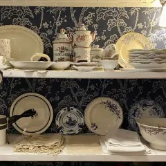 Étagères remplies de vaisselle ancienne, dont des tasses, des assiettes et des bols, avec un motif floral bleu sur fond sombre. Une étiquette mentionne "Porcelaine de Paris signature".