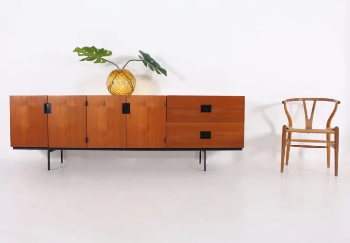 Buffet en teck par Cees Braakman, avec un vase en verre texturé et une feuille de Monstera. Une chaise Wishbone est placée à droite.