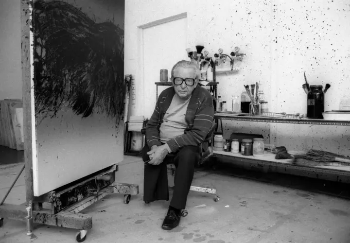 Hartung, peintre, pose dans son atelier, devant une de ses toiles abstraites. L'espace est encombré de matériel artistique.