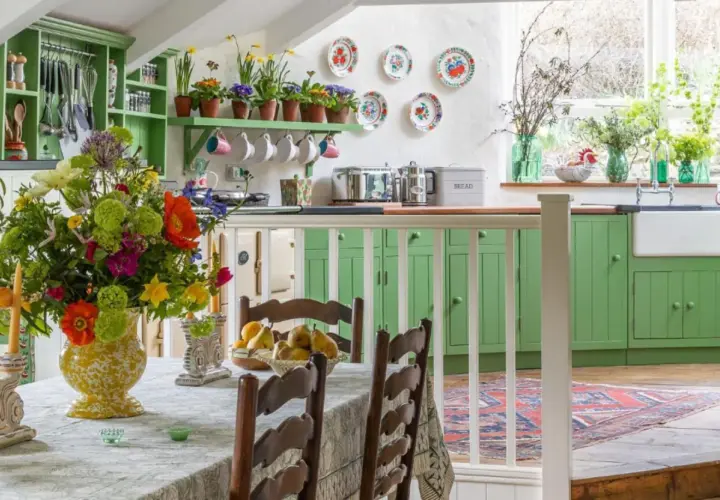 Une cuisine lumineuse avec des armoires vertes vives, un grand bouquet de fleurs colorées sur la table, et une décoration style cottage ou campagne chic.