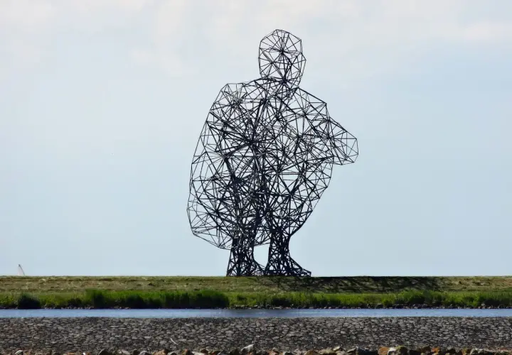 Sculpture d'Antony Gormley, une silhouette humaine filaire accroupie, se dresse sur une digue herbeuse, surplombant un canal.