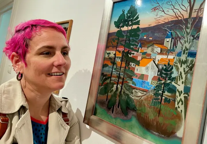 Une femme aux cheveux roses admire un tableau d'Étienne Cournault représentant un village pittoresque sous un ciel coloré.