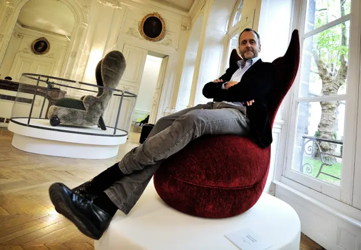 Hubert Le Gall, designer, pose dans un fauteuil rouge audacieux, entouré d'œuvres d'art dans un salon élégant.