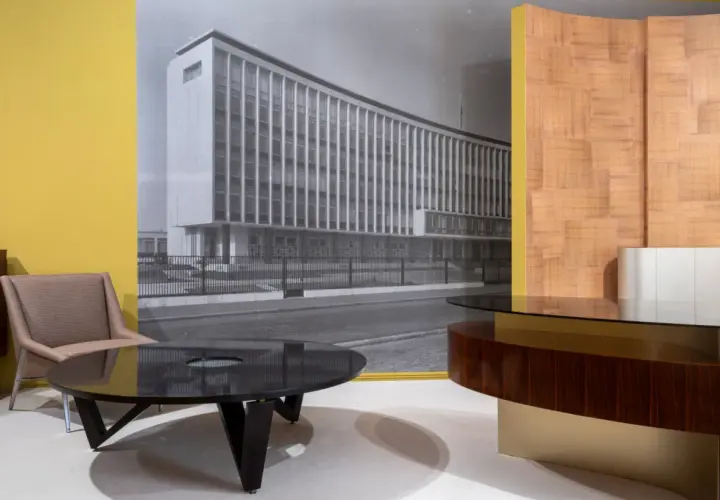 Un fauteuil et une table basse devant un mur jaune, avec une photo en noir et blanc d'un bâtiment moderne, rappelant le style de Maxime old.