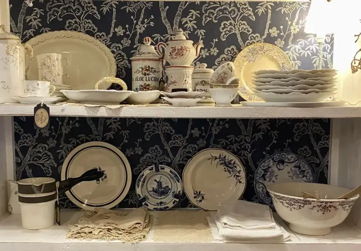 Étagères remplies de vaisselle ancienne, dont des tasses, des assiettes et des bols, avec un motif floral bleu sur fond sombre. Une étiquette mentionne "Porcelaine de Paris signature".