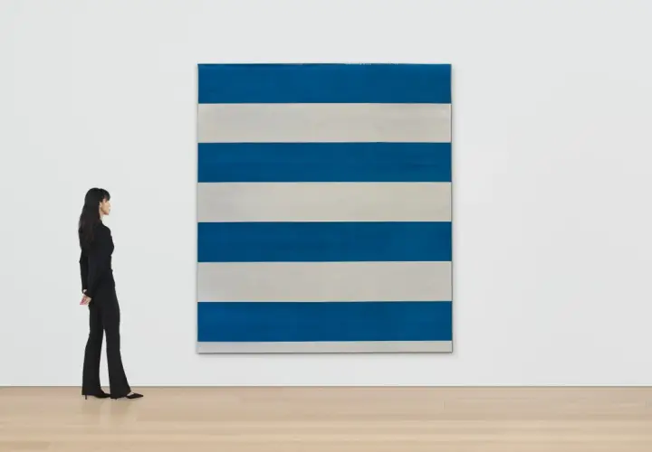 Une femme observe une œuvre de Michel Parmentier, une grande toile rayée de bleu et de blanc, dans une galerie d'art.