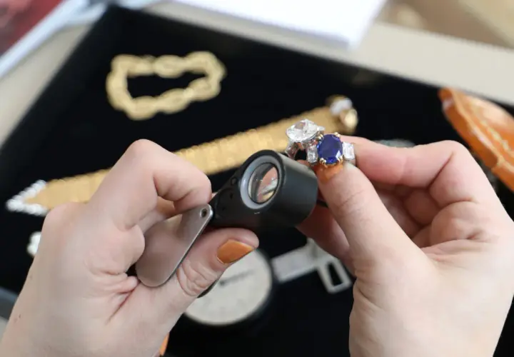 Maintenant, où faire expertiser des bijoux gratuitement ? Une main examine une bague saphir et diamant avec une loupe.