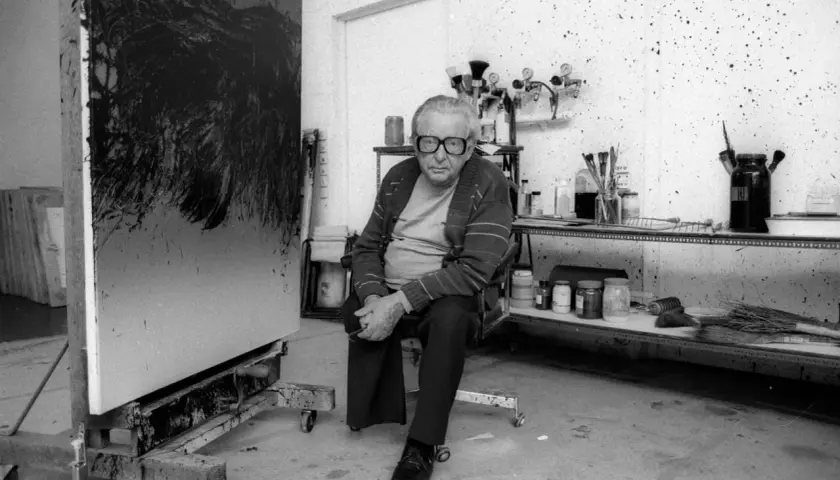 Hartung, peintre, pose dans son atelier, devant une de ses toiles abstraites. L'espace est encombré de matériel artistique.