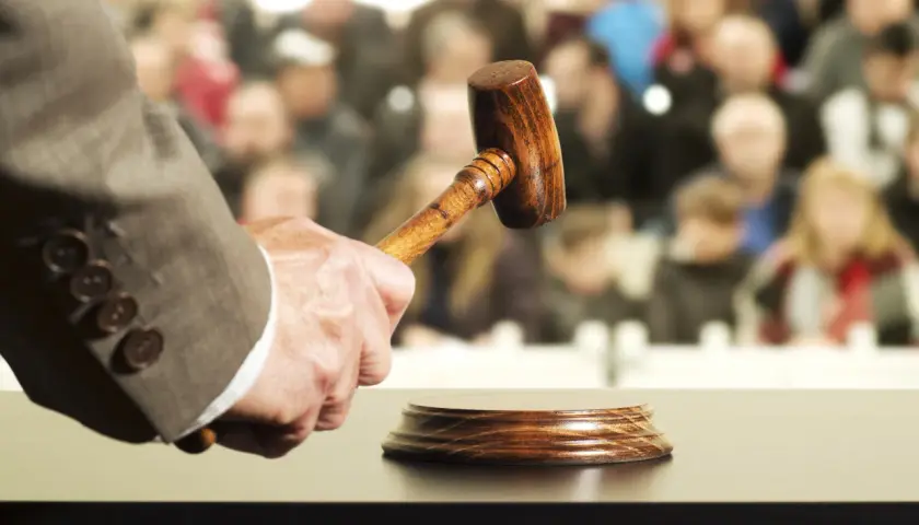 Un commissaire-priseur tient un maillet, prêt à frapper le bloc. La foule attend, espérant acquérir un objet convoité, peut-être au-delà du prix de réserve enchère.