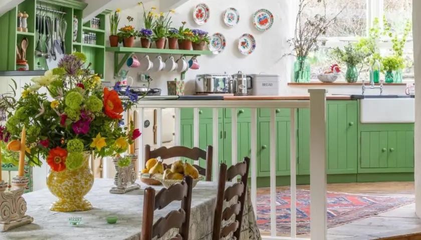 Une cuisine lumineuse avec des armoires vertes vives, un grand bouquet de fleurs colorées sur la table, et une décoration style cottage ou campagne chic.