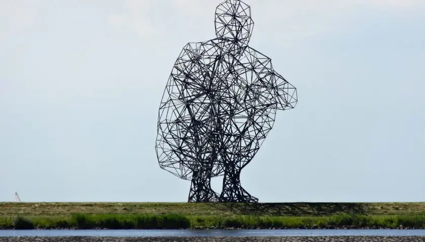 Sculpture d'Antony Gormley, une silhouette humaine filaire accroupie, se dresse sur une digue herbeuse, surplombant un canal.