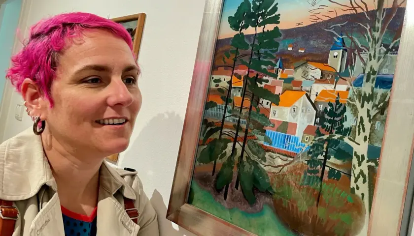 Une femme aux cheveux roses admire un tableau d'Étienne Cournault représentant un village pittoresque sous un ciel coloré.