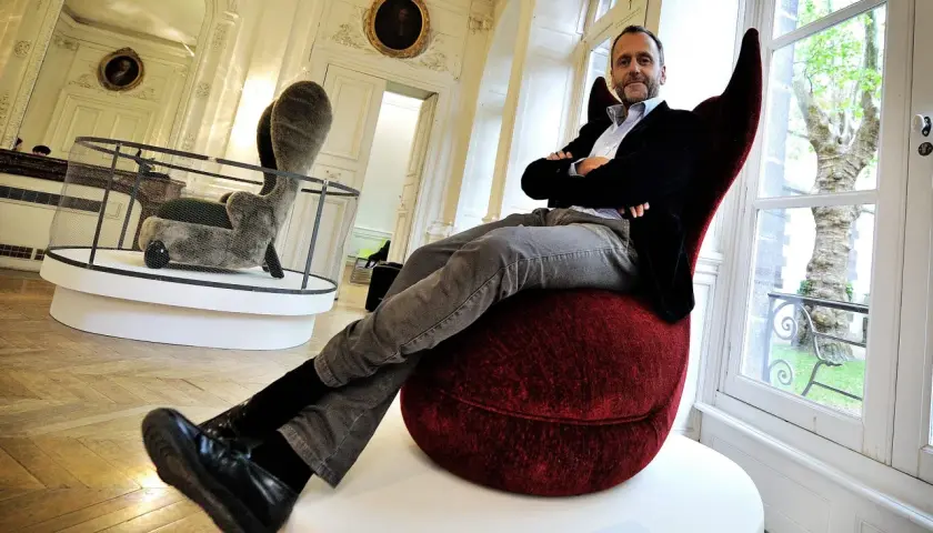 Hubert Le Gall, designer, pose dans un fauteuil rouge audacieux, entouré d'œuvres d'art dans un salon élégant.
