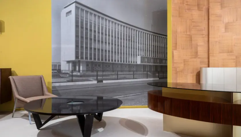 Un fauteuil et une table basse devant un mur jaune, avec une photo en noir et blanc d'un bâtiment moderne, rappelant le style de Maxime old.