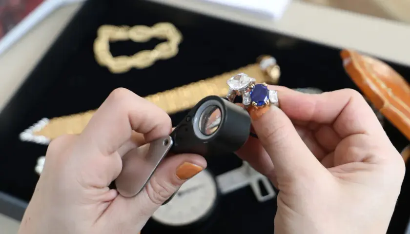 Maintenant, où faire expertiser des bijoux gratuitement ? Une main examine une bague saphir et diamant avec une loupe.
