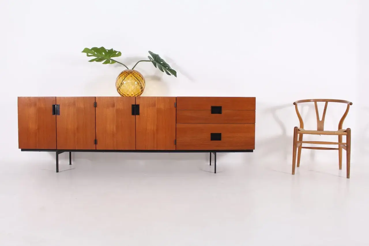 Buffet en teck par Cees Braakman, avec un vase en verre texturé et une feuille de Monstera. Une chaise Wishbone est placée à droite.