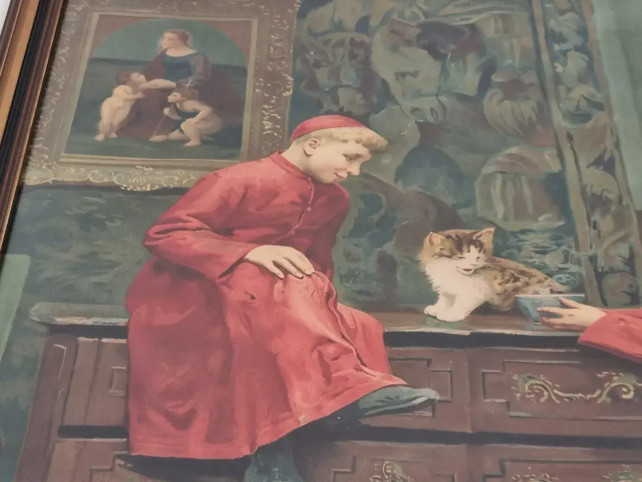 Un jeune chanoine moreau, en robe rouge, observe un chaton joueur. Derrière lui, un tableau représente une scène biblique.