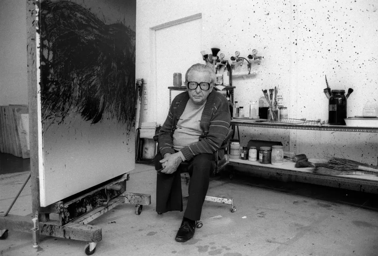 Hartung, peintre, pose dans son atelier, devant une de ses toiles abstraites. L'espace est encombré de matériel artistique.