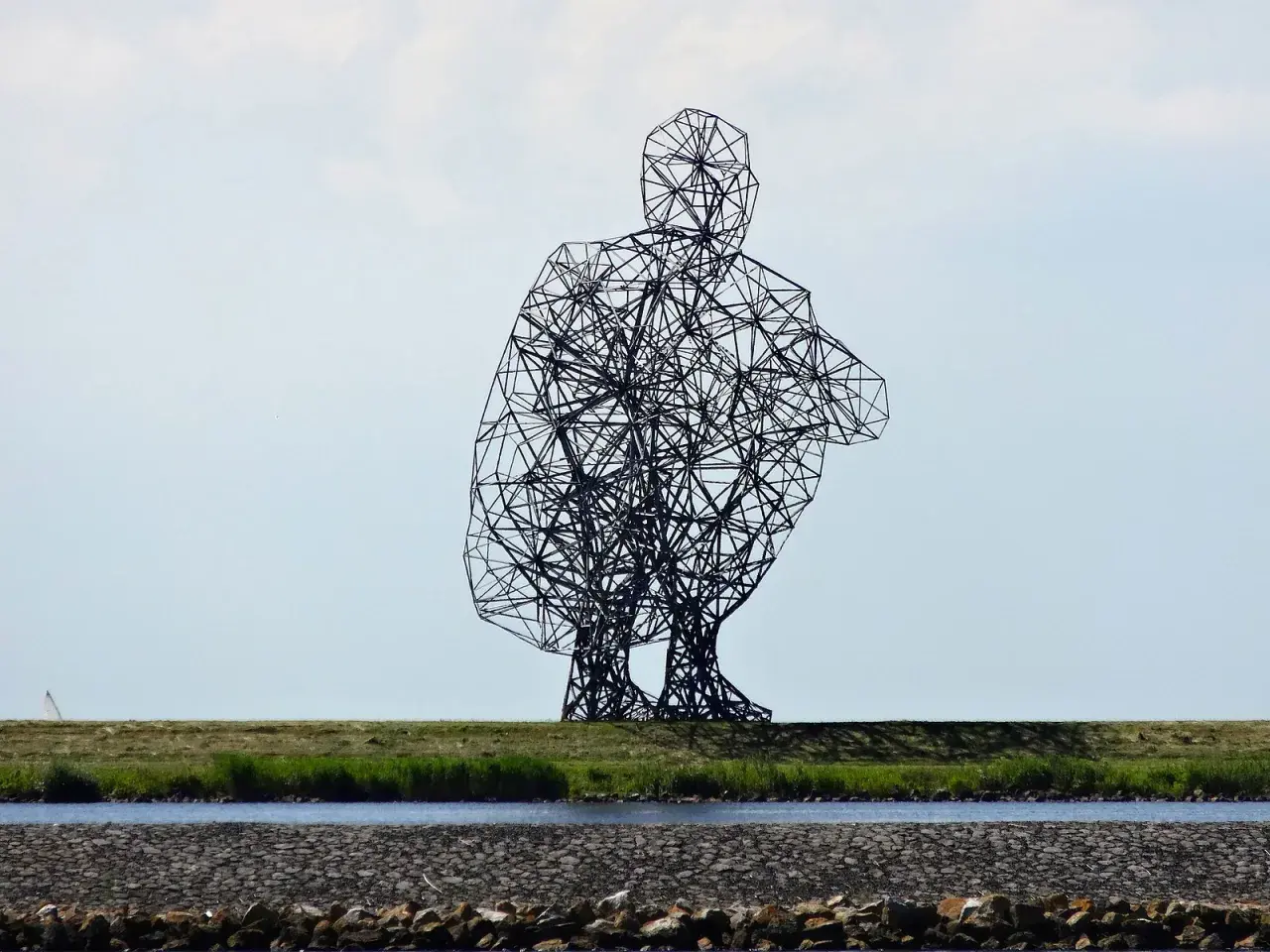 Sculpture d'Antony Gormley, une silhouette humaine filaire accroupie, se dresse sur une digue herbeuse, surplombant un canal.