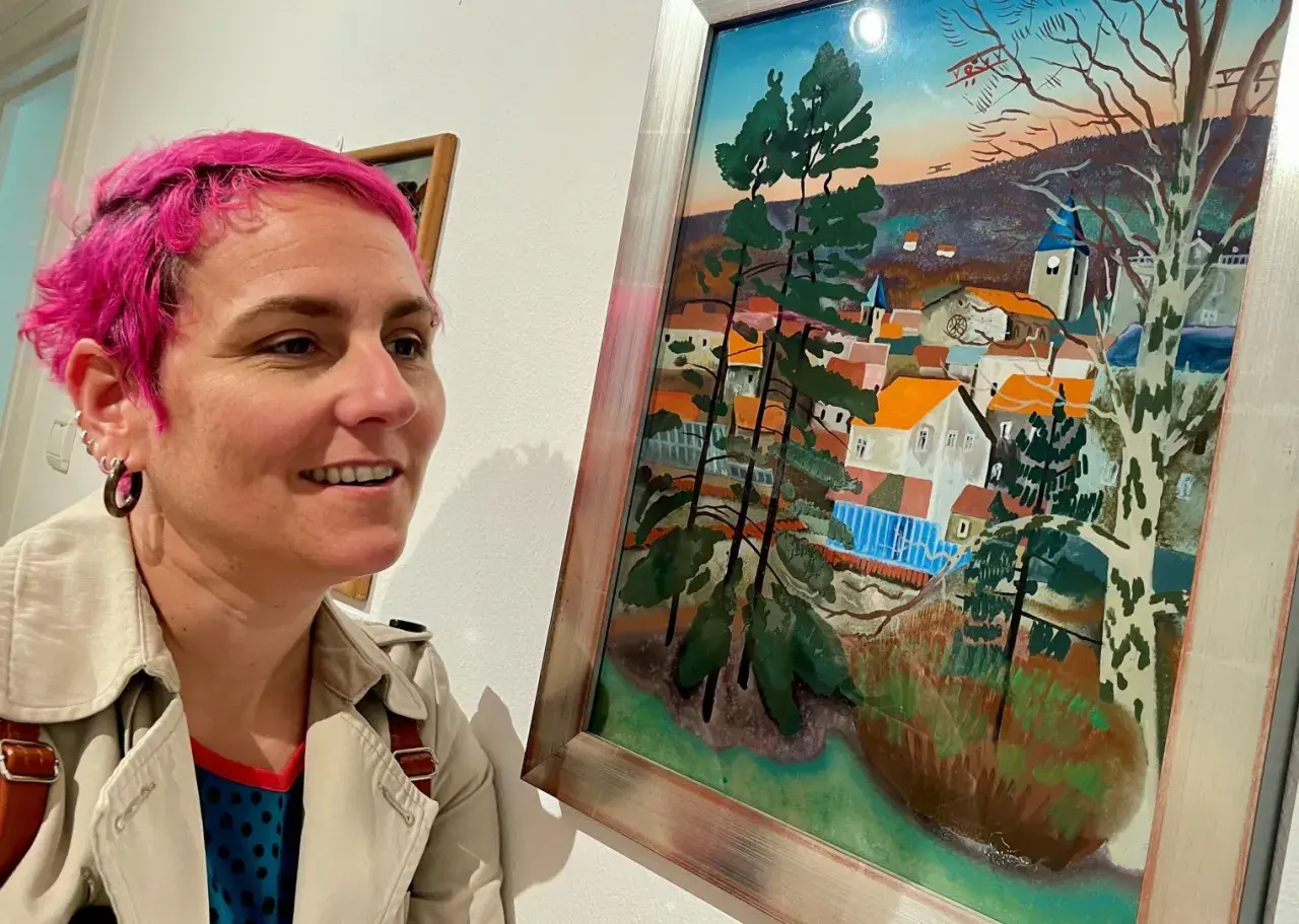 Une femme aux cheveux roses admire un tableau d'Étienne Cournault représentant un village pittoresque sous un ciel coloré.