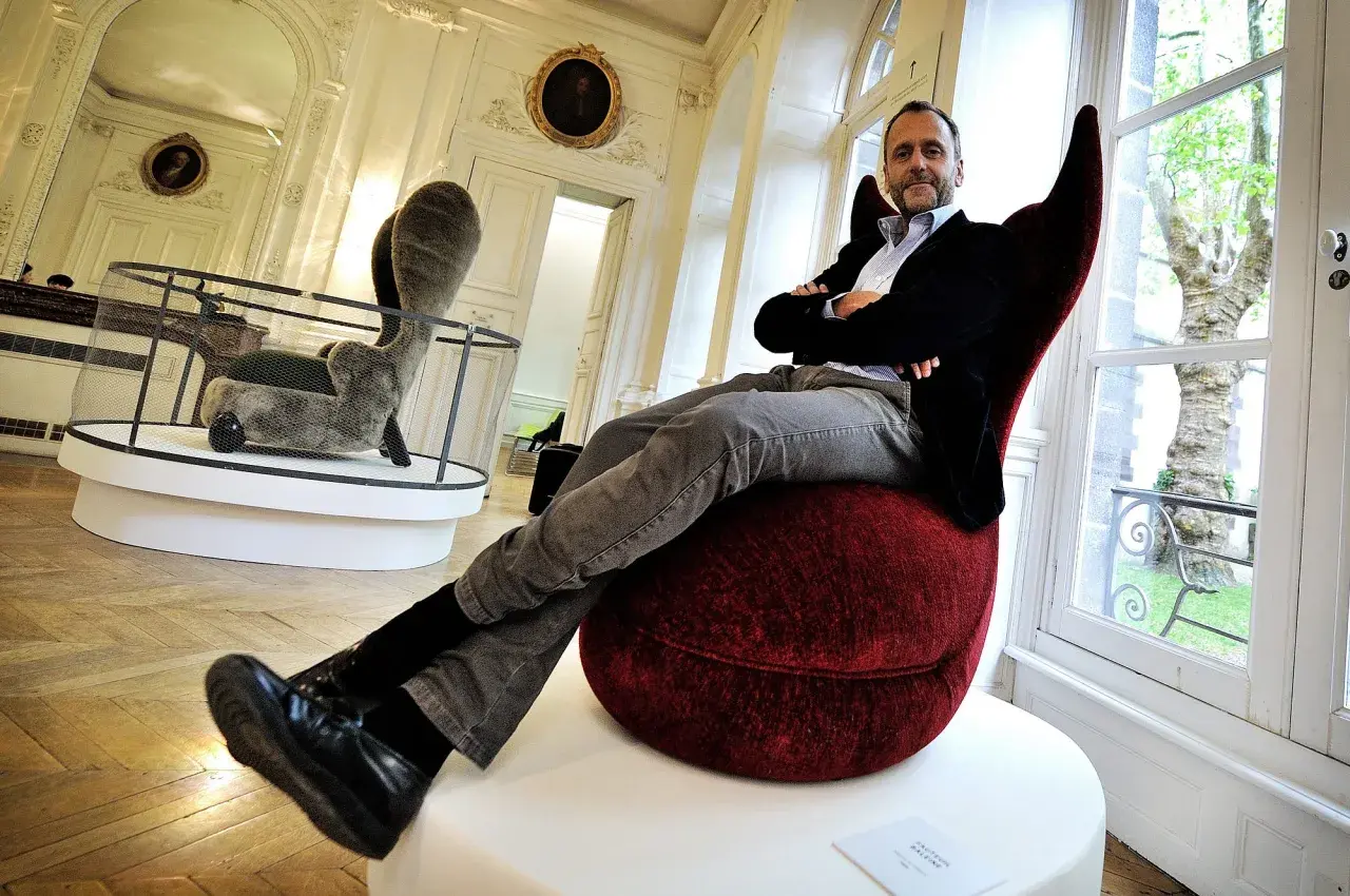 Hubert Le Gall, designer, pose dans un fauteuil rouge audacieux, entouré d'œuvres d'art dans un salon élégant.