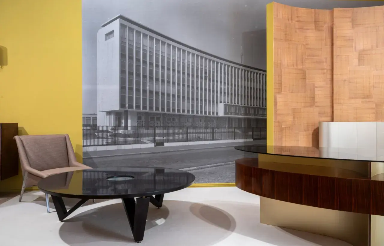 Un fauteuil et une table basse devant un mur jaune, avec une photo en noir et blanc d'un bâtiment moderne, rappelant le style de Maxime old.