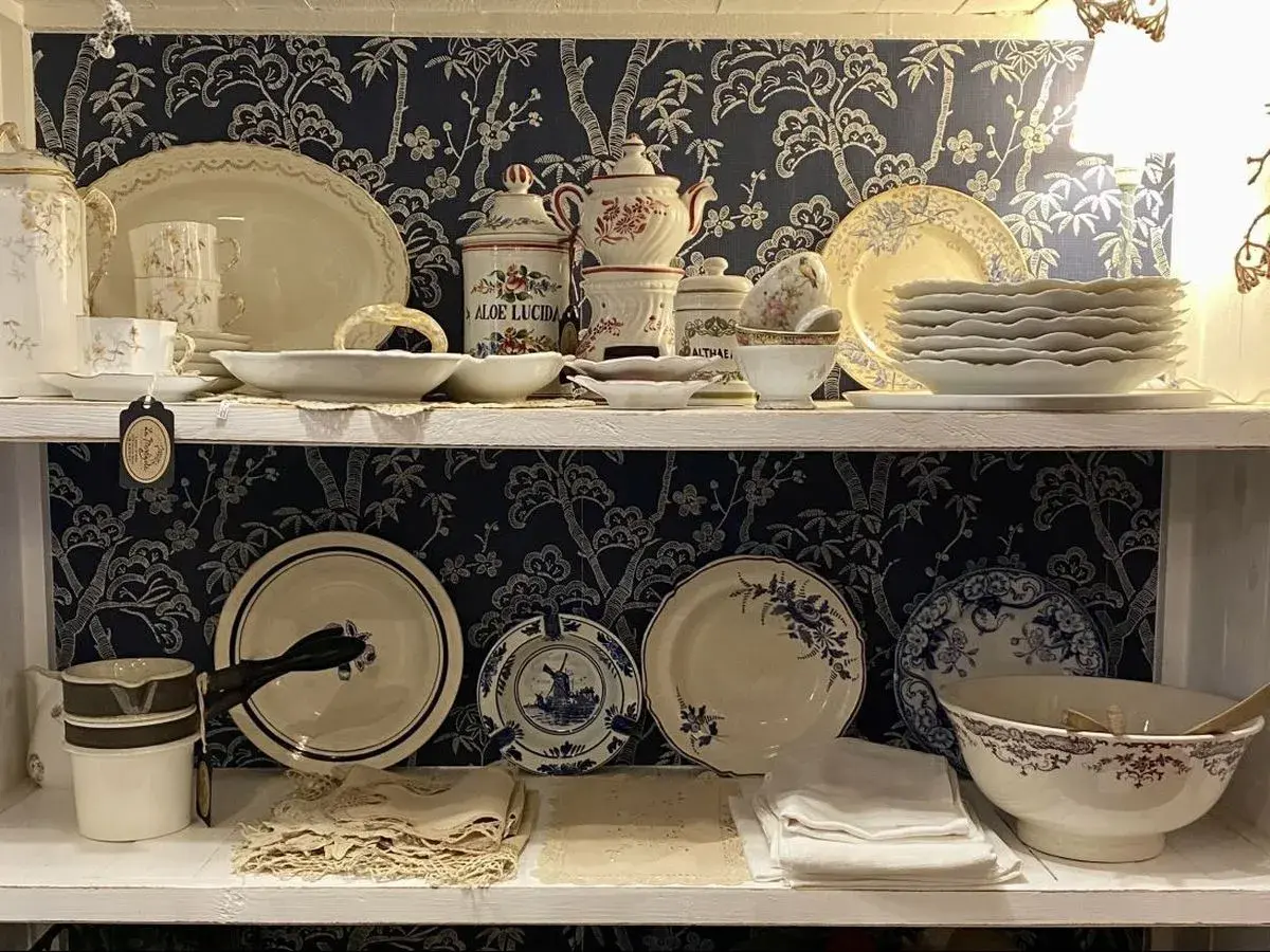 Étagères remplies de vaisselle ancienne, dont des tasses, des assiettes et des bols, avec un motif floral bleu sur fond sombre. Une étiquette mentionne "Porcelaine de Paris signature".