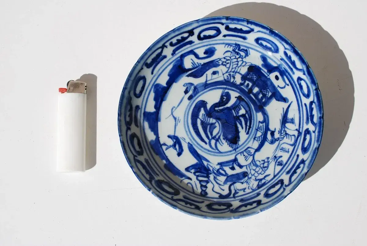 Assiette chinoise ancienne signature, décorée de motifs bleus sur fond blanc, avec un briquet à côté pour l'échelle.
