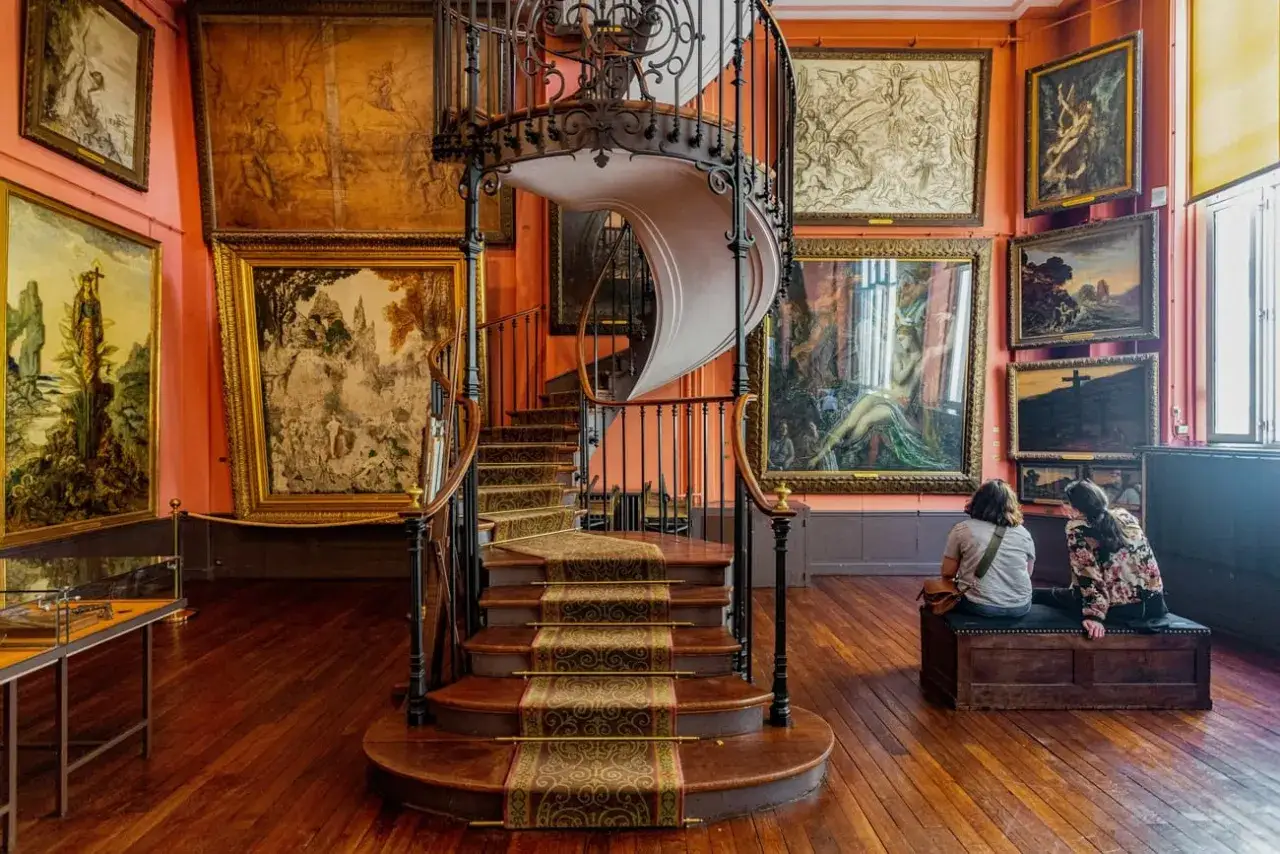 Escalier en colimaçon dans un musée, entouré d'œuvres de Gustave Moreau. Deux visiteuses contemplent les toiles.