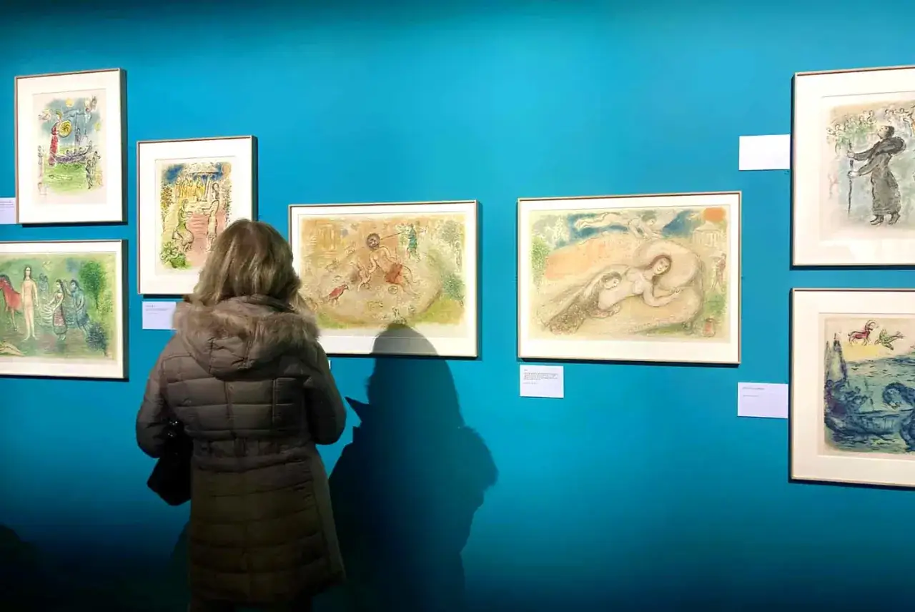 Une femme admire des &oelig;uvres d'art de Marc Chagall sur un mur bleu vif. Les tableaux repr&eacute;sentent des sc&egrave;nes oniriques et color&eacute;es.