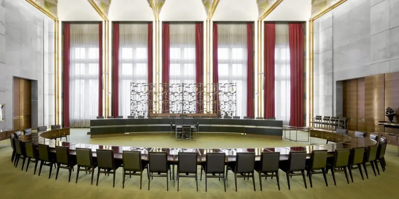 Salle de conférence majestueuse avec une grande table ovale, des sièges en cuir et des rideaux rouges. Un décor d'époque, peut-être pour une réunion de Maxime Old.