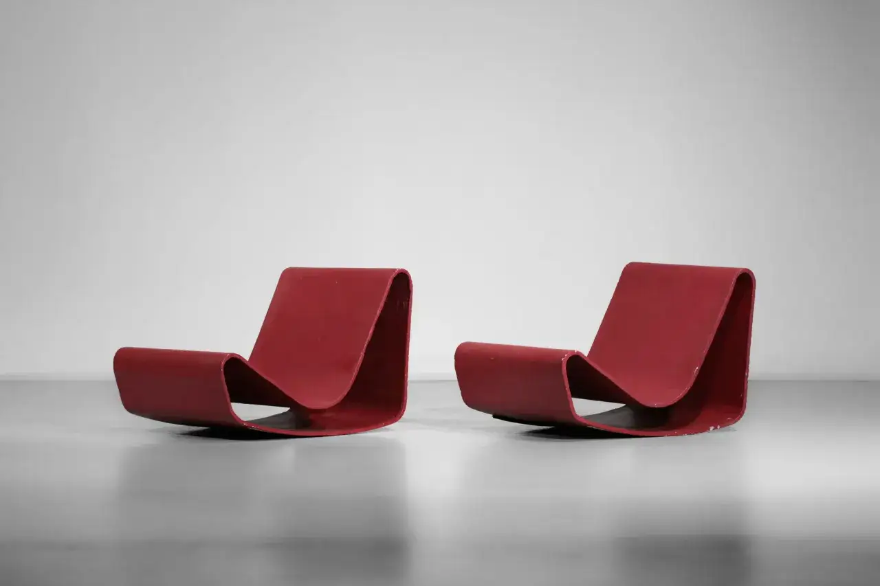 Deux fauteuils bas rouges iconiques, cr&eacute;&eacute;s par Willy Guhl, aux formes courbes et organiques, pos&eacute;s sur un sol gris.